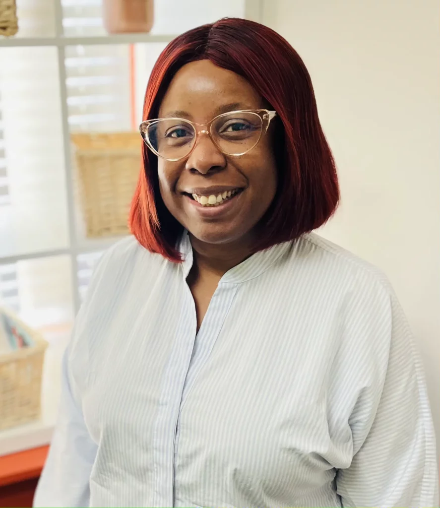 Temi Sokunbi with red hair and glasses smiles in front of a window physiotherapy.