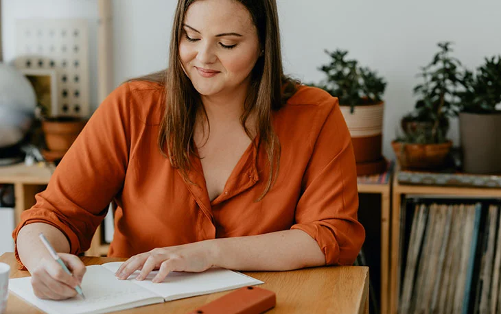 A woman diligently focused at a table writing in a notebook, prioritizing her back health as part of her New Year's resolutions.